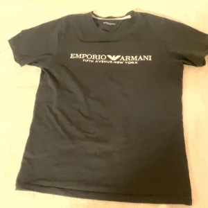 Svart Emporio Armani t-shirt - Svart t-shirt från Emporio Armani med vit logotyp och text 'Fifth Avenue New York' på bröstet. Klassisk rund hals och korta ärmar. Stilren design som passar perfekt till jeans eller shorts. Materialet känns mjukt och bekvämt mot huden.