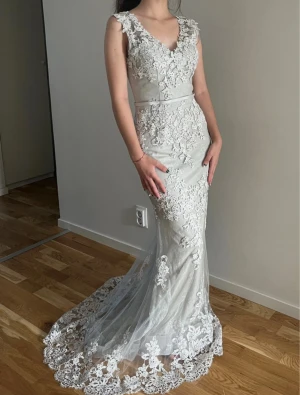 Vit lång spetsklänning med v-ringning - Elegant vit långklänning med v-ringning och vackra spetsdetaljer över hela klänningen. Klänningen har ärmlös design och markerad midja med ett smalt bälte. Nederdelen har ett släp med extra mycket spets, vilket ger en lyxig känsla.