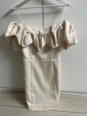 Champagnefärgad kortklänning med volang - Supersnygg kortklänning i champagnefärg med breda volanger över axlarna och smala axelband. Klänningen har en figurnära passform och är tillverkad i ett slätt, mjukt material som ger en lyxig känsla. Perfekt för dig som vill sticka ut med en unik och trendig look.