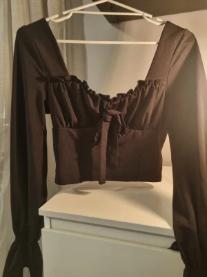 Svart croppad blus med snörning - Säljer en svart croppad blus med långa ärmar och fyrkantig urringning. Toppen har volangkant upptill, snörning framtill och dragkedja i ryggen.💞