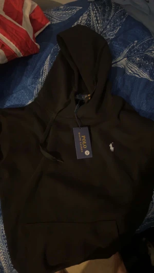 Ralph lauren - Det är en svart huvtröja från Polo Ralph Lauren. På bröstet finns det klassiska lilla broderade logotypsmotivet med en ryttare och häst i vitt. Huvan har svarta dragsnören och framtill sitter en mörk, rektangulär prislapp med guldtext där det står POLO RALPH LAUREN samt storleken “S”. Huvtröjan ligger utlagd på ett blått täcke med vita mönster. Den ser ny och oanvänd ut med taggen fortfarande kvar.