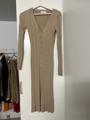 Beige ribbad långklänning med knappar - Säljer en beige ribbad långklänning med v-ringning och långa ärmar. Klänningen har knappar hela vägen framtill och är figurnära i modellen. Perfekt för dig som gillar stilrena och enkla plagg med snygga detaljer.