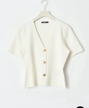 Vit v-ringad topp från Gina Tricot - Säljer en stilren vit topp från Gina Tricot med v-ringning och korta ärmar. Toppen har tre snygga guldfärgade knappar framtill och är lätt croppad i modellen. Perfekt för dig som gillar enkel och fräsch stil. Använd 1 gång