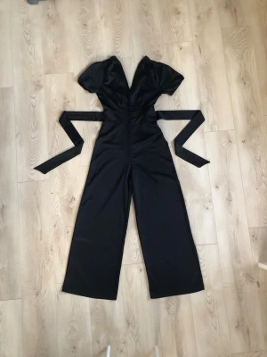 Satin jumpsuit  - Festlig svart jumpsuit med korta puffärmar, djup v-ringning och klädda knappar framtill. Knytband i midjan som framhäver figuren. Vida ben och mjukt, glansigt tyg som ger en elegant känsla. Mycket snygg och samtidigt bekväm.