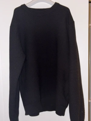 Svart stickad tröja  - Svart stickad tröja   med rund hals och långärmad design. Tröjan har en diskret textur i materialet och ribbade muddar vid hals, ärmslut och nederkant. Perfekt för dig som gillar stilrena och klassiska plagg med en exklusiv touch. Den är inte sol blekt 