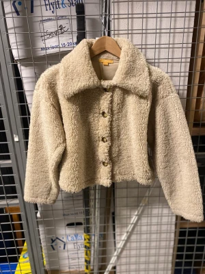 Beige teddyjacka med knappar - Mysig beige teddyjacka med krage och knappar framtill. Jackan är croppad och har långärmade ärmar, perfekt för att ge en chill och trendig vibe. Materialet är fluffigt och ger en skön känsla, passar dig som gillar streetstyle och vill ha något som sticker ut.
