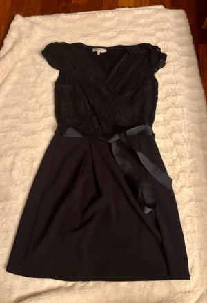 Mörkblå spetsklänning - Supersnygg mörkblå klänning från Morgan med spets upptill, omlott och v-ringning. Midjan markeras av ett satinband som knyts till rosett. Kjolen är enkel och stilren, medan överdelen har blommig spets och korta ärmar. Perfekt för dig som vill ha festlig och elegant look.