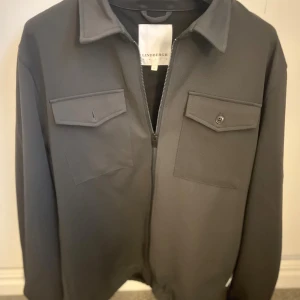 Grå overshirt från Lindbergh - Svart overshirt från Lindbergh med klassisk krage, två stora bröstfickor med lock och knapp, samt dragkedja framtill. Jackan har en clean och stilren look, perfekt för lager-på-lager. Materialet är mjukt och känns lätt mot kroppen. Passformen är normal. Storlek L. Skriv vid frågor! 🙌✅