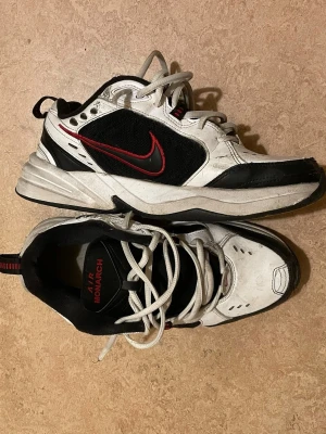 Nike Air Monarch sneakers svart/vit - Nike Air Monarch sneakers i svart och vitt med röda detaljer och klassisk swoosh på sidan. Skorna har snörning, platt sula och en sportig, chunky design. Materialet är en mix av syntet och skinn, perfekt för dig som gillar retrostil och komfort.