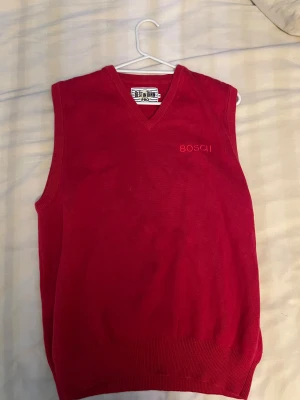 Röd stickad väst från Best In Town - Snygg röd stickad väst från Best In Town med v-ringad hals och broderad BOSCI-text på bröstet. Perfekt att slänga över en skjorta för en clean och klassisk look. Ribbad kant nedtill och vid ärmhålen för extra stilpoäng.