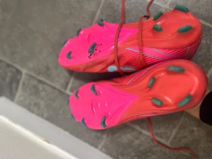 Röda Nike fotbollsskor med blå swoosh - Säljer ett par riktigt snygga röda Nike fotbollsskor påse till skorna hänger med med ljusblå swoosh på sidorna. Skorna har snörning, platt sula med gröna dobbar och är tillverkade i syntetmaterial. Perfekta för dig som vill sticka ut på planen med stilren design och grym passform.