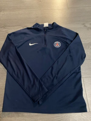 PSG Nike Dri-Fit träningströja XL - Mörkblå långärmad träningströja från Nike med PSG-logga på bröstet och vit swoosh. Tillverkad i Dri-Fit material som andas, halv dragkedja framtill och stilren design. Perfekt för fotbollsträning eller sportiga dagar.