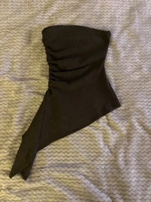 Mörk grön asymmetrisk tubtopp - Säljer en mörk grön tubtopp med unik asymmetrisk design och rynkade detaljer på ena sidan. Toppen är axelbandslös och har en cool, modern form som sticker ut. Perfekt för dig som vill ha något annorlunda och trendigt i garderoben.