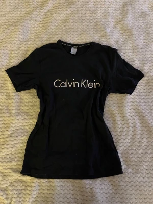 Svart Calvin Klein t-shirt - Svart t-shirt från Calvin Klein med klassisk logga tryckt i vitt över bröstet. T-shirten har rund halsringning och korta ärmar. Tillverkad i mjuk bomull för en skön och avslappnad look.