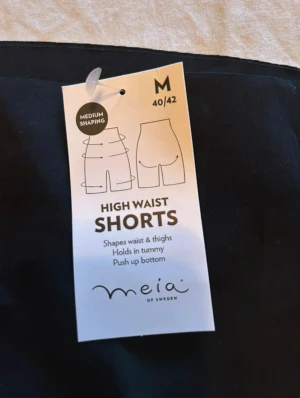 Svarta shaping shorts från Meia - Svarta high waist shaping shorts från Meia of Sweden i storlek M. De har en riktigt hög midja som formar midja och lår, håller in magen och ger push up-effekt för rumpan. Perfekta under klänning eller kjol för en slät siluett.