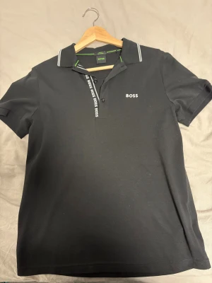 Svart Boss piké med vit logga - Svart pikétröja från Boss med vit broderad logga på bröstet och vit text längs knappslån. Kragen har vita ränder och insidan är vit. Grön detalj på insidan av kragen. Tillverkad i mjuk pima bomull, kortärmad och slim fit för en clean look.