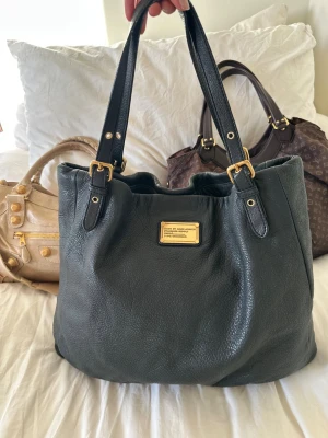 Snygg Shopgirl Tote från Marc Jacobs! - Snyggaste svarta läderväskan i äkta läder från Marc by Marc Jacobs. Den ikoniska modellen Q Classic Shopgirl Tote! En populär och eftertraktad väska med gulddetaljer och dubbla handtag. Lädret är mjukt, slitstarkt och i fint skick.   Det här är en klassisk och rymlig modell som inte längre tillverkas, vilket gör den extra eftertraktad. Passar perfekt för dig som vill ha en stilren väska med hög kvalitet och riktigt bra andrahandsvärde. Rymmer både dator och gymkläder och allt du behöver!🤍