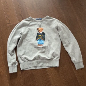 Grå sweatshirt Polo Bear Ralph Lauren - Grå sweatshirt från Polo Ralph Lauren med ikoniskt Polo Bear-tryck på bröstet. Rund hals, ribbade muddar och nederkant. Mjuk bomullskvalitet och långärmad modell. Perfekt för dig som gillar klassisk streetstyle med en twist. Nypris 1800