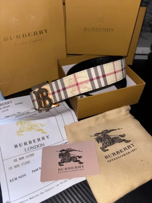 Burberry vändbart bälte - Snyggt vändbart Burberry-bälte i kalvskinn med klassiskt rutmönster i beige, svart och rött på ena sidan och svart präglad läder på andra. Metallspänne med stort B-monogram i guld. Kommer med originalförpackning, dustbag och certifikat.Den bältet som du ser på bilden är för show. Jag fraktar ut helt nya fortfarande inplastad!!!