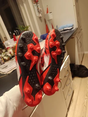 Röda och svarta fotbollsskor med mönster - Säljer ett par fotbollsskor i rött, svart och vitt med coolt grafiskt mönster och snörning. Skorna har rund tå och platt sula med röda dobbar för gräsplan. Insidan är svart och vit, och ovandelen är tillverkad i syntetmaterial för lätt känsla på foten.