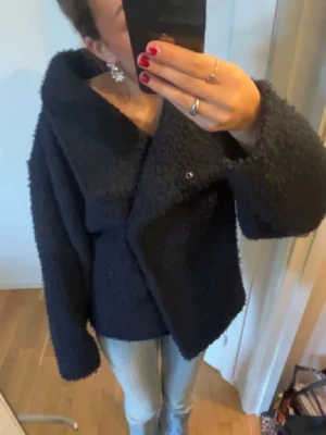 Mörkblå kappa  - Säljer en svart teddyjacka med stor, asymmetrisk krage och breda ärmar. Jackan har en loose passform och stängs med knappar framtill. Perfekt för dig som gillar en chill och trendig look under höst och vinter.