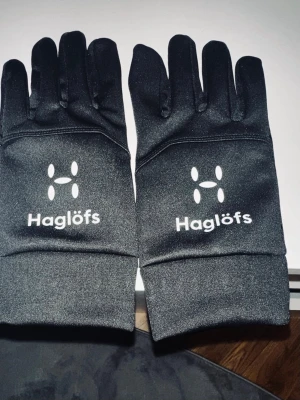 Haglöfs Handskar  - Svarta sporthandskar från Haglöfs med vit logga på ovansidan. Handskarna har greppdetaljer på insidan av fingrarna och handflatan för extra fäste. Tillverkade i syntetmaterial som är smidigt och stretchigt, perfekta för utomhusaktiviteter under kyliga dagar.