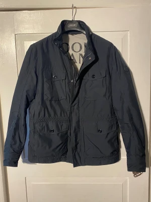 Fieldjacka / fieldjacket JOOP! Jeans - Mörkblå fieldjacka från joop jeans. Vädermotståndeligt yttre skal med fodrat inre. Försluten med dragkedja samt en knappförsluten windflap som täcker den. Fyra fickor fram med knappförslutning samt två dolda sidofickor bakom de nedre. Ståkrage med dragkedja samt en justerbar midja. En perfekt allvädersjacka som du kan ha långt in i de kallare av vinterdagarna utan att frysa. Använd några enstaka gånger precis vid nyköp! Om du undrar över någonting så är det bara att fråga! //Hugo