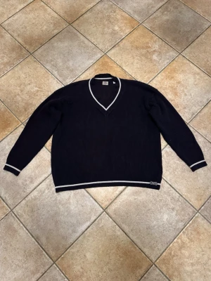 Mörkblå stickad tröja från Levi's XL - Snygg mörkblå stickad tröja från Levi's i storlek XL. Tröjan har v-ringad hals och vita ränder vid hals, ärmslut och nederkant. Ribbad struktur och klassisk Levi's-logga nertill. Perfekt för dig som gillar stilrena och sportiga vibbar.