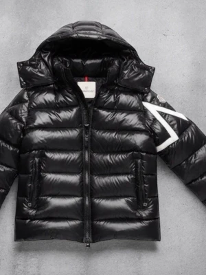 Svart Moncler Maya  - Säljer en svart pufferjacka från Moncler med glansig finish och stor huva. Jackan har en vit detalj på ena ärmen, två fickor med dragkedja och en kraftig frontdragkedja. Perfekt för kalla dagar och har en riktigt snygg streetstil.