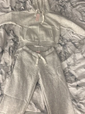 Ljusgrå mjukisbyxor med snörning tröjan (helt oanvänd) - Supersköna ljusgrå mjukisbyxor från Ginatricot i mjuk bomullsmix. Byxorna har dragsko i midjan, ribbade muddar och en bakficka med broderad detalj. Perfekta för chill dagar eller när du vill vara extra bekväm.