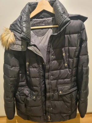 Svart dunjacka Marc O'Polo med huva  - Säljer en svart dunjacka från Marc O'Polo med quiltad design och stor huva med fuskpäls. Jackan har flera fickor med tryckknappar, dragkedja framtill och ribbade detaljer vid kragen. 