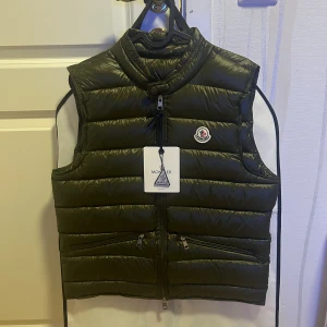 Moncler Väst - Snygg olivgrön dunväst från Moncler med quiltad design och klassisk logga på bröstet. Västen har hög krage, dragkedja framtill och två snedställda fickor med dragkedja. Perfekt för lager-på-lager och streetstyle. OBS sista bilden pytte liten fläck som kan gå bort i tvätt