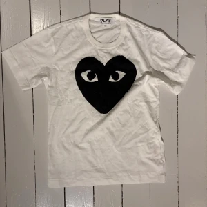 Comme des Garçons t-shirt  - Säljer en vit t-shirt från Comme des Garçons Play i storlek S. Framsidan har ett stort svart hjärta med ögon, vilket ger en unik och edgy vibe. T-shirten är kortärmad och gjord i mjuk bomull med rund hals. Perfekt för dig som gillar streetwear och grafiska prints.