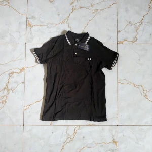 Svart piké från Fred Perry - Säljer en svart piké från Fred Perry med vit broderad logga på bröstet och vita ränder på krage och ärmslut. Klassisk polokrage med två knappar framtill. Perfekt för dig som gillar stilrena och sportiga plagg.