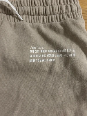 Beige shorts  - Snygga beige shorts med vitt tryck på framsidan och texten 'new york'. De har elastisk midja med vit snörning för skön passform. Materialet är mjukt och känns som bomull, perfekt för chill dagar. Passar dig som gillar enkel och clean stil.