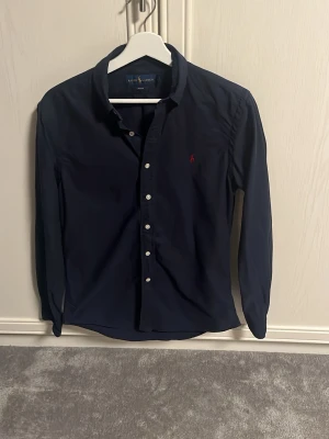 Ralph lauren skjorta Intressekoll! - INTRESSEKOLL! Hur mycket skulle ni kunna betala för denna. Säljer en mörkblå slim fit skjorta från Ralph Lauren med klassisk krage och vita knappar. Skjortan har lång ärm och den ikoniska röda logotypen broderad på bröstet. Perfekt till jeans eller chinos för en clean och stilren look.