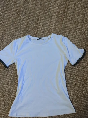 Vit t-shirt med glittriga ärmslut från Zara - Snygg vit t-shirt från Zara i storlek M. T-shirten har korta ärmar med glittriga svarta detaljer vid ärmsluten som ger en extra touch. Materialet känns mjukt och skönt mot huden, perfekt för dig som gillar stilrena plagg med en twist.