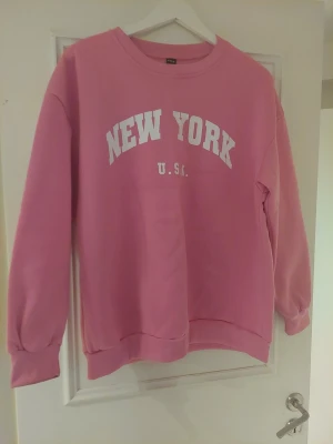 Rosa sweatshirt med New York-tryck - Cool rosa sweatshirt från VRS med vit text 'NEW YORK U.S.A.' på bröstet. Tröjan har rund hals, långa ärmar och ribbade muddar vid ärmslut och nederkant. Perfekt för dig som gillar streetstyle och vill sticka ut med färg.