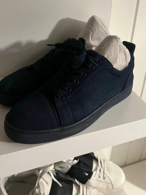 Mörkblå sneakers från Louboutin - Snygga mörkblå sneakers från Louboutin med klassisk röd sula och rund tå. Skorna har snörning och är tillverkade i mocka för en lyxig känsla. Perfekt för dig som vill sticka ut med exklusiva detaljer och stilren design.