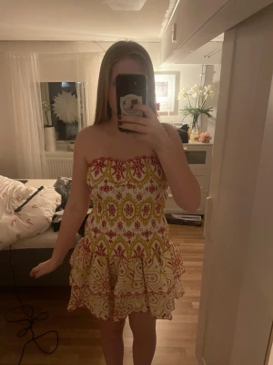 Mönstrad axelbandslös kortklänning - Superfin axelbandslös kortklänning med volangkant och färgglatt mönster i rött, gult och vitt. Klänningen har en smockad överdel och luftig kjol med två volanger. Perfekt för varma dagar och somriga vibes!