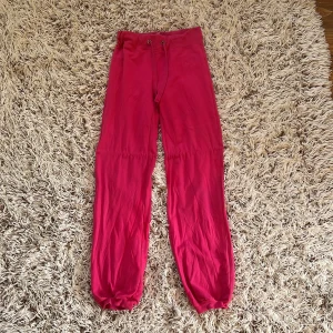 Rosa mjukisbyxor från H&M Sport  - Säljer ett par mjukisbyxor från H&M Sport i en stark rosa färg.  Byxorna har snörning i midjan (Tyvärr saknar ena delen av snöret sin metal bit, se bilderna), ribbade muddar vid bensluten och är tillverkade i mjukt bomullsmaterial.  Perfekta för chill eller träning. Finns broderad logga på sidan. Storlek XS