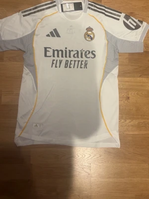 Real Madrid adidas matchtröja vit - Säljer en officiell Real Madrid matchtröja från adidas i vit med grå och gula detaljer. Tröjan har klubbmärket på bröstet, sponsortryck och klassiska adidas-ränder på axlarna. Perfekt för dig som älskar fotboll och vill ha en snygg tröja med modern design.