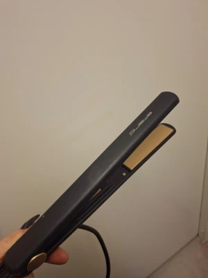 Svart plattång från Emerio - Snygg svart plattång från Emerio med guldiga keramiska plattor och smidig design. Perfekt för att styla håret och skapa både raka och vågiga looks. Har en enkel knapp för temperaturjustering och lång sladd för flexibel användning.