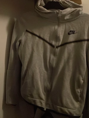 Grå nike tech size xs - Nike vindjacka i ljusgrått med huva och hel dragkedja. Svarta och beige detaljer längs bröstet och ärmarna, samt Nike-logga på framsidan. Jackan har fickor med dragkedja och en clean, sportig look. Perfekt för dig som gillar streetwear.