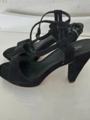 Svarta sandaler med klack och snörning - Säljer ett par svarta sandaler från Åhléns med hög klack och snygga remmar som går runt foten. Skorna har öppen tå och spänne vid ankeln. Materialet är mocka och sulan är platt framtill. Perfekta för dig som gillar stilrena och trendiga skor.