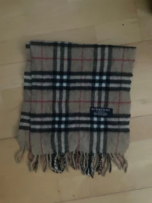 Rutig beige halsduk från Burberry - Säljer en klassisk halsduk från Burberry i beige med rutigt mönster i svart, vitt och rött. Halsduken är tillverkad i 100% lammull och har fransar i kanterna. Perfekt accessoar för att lyfta din outfit med lite lyxig London-vibe. Längd:125 bredd 26