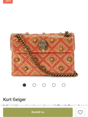 Orange axelväska från Kurt Geiger - Säljer en orange axelväska från Kurt Geiger med glittriga detaljer och kedjerem. Väskan har ett rutigt mönster med guldiga nitar och en unik dekoration på locket. Perfekt för dig som vill sticka ut med en färgglad och trendig accessoar. Köpt för 2400