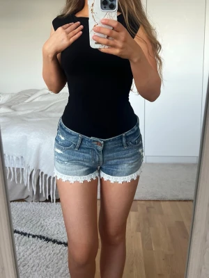 Blå jeansshorts med spets från Hollister 🩵 - Säljer ett par blå jeansshorts från Hollister med låg midja och snygga slitningar. Shortsen har vit spetskant med blommönster längst ner 🩵
