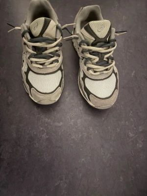 Gråa Asics sneakers - Säljer ett par gråa Asics sneakers med vita och mörkgrå detaljer. Skorna har en sportig look med meshpaneler för bra ventilation och partier i mocka. Klassisk snörning och rund tå. Perfekta för dig som gillar stilrena och bekväma sneakers.