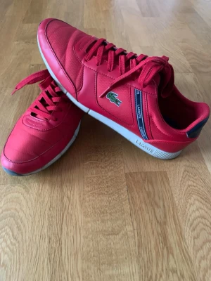 Röda sneakers från Lacoste - Snygga röda sneakers från Lacoste med klassisk krokodillogo på sidan och mörkblå detaljer. Skorna har snörning, rund tå och platt sula. Ovandelen är i syntetmaterial med textilinslag och vita detaljer på sulan. Perfekt för dig som gillar sportig stil och färg.
